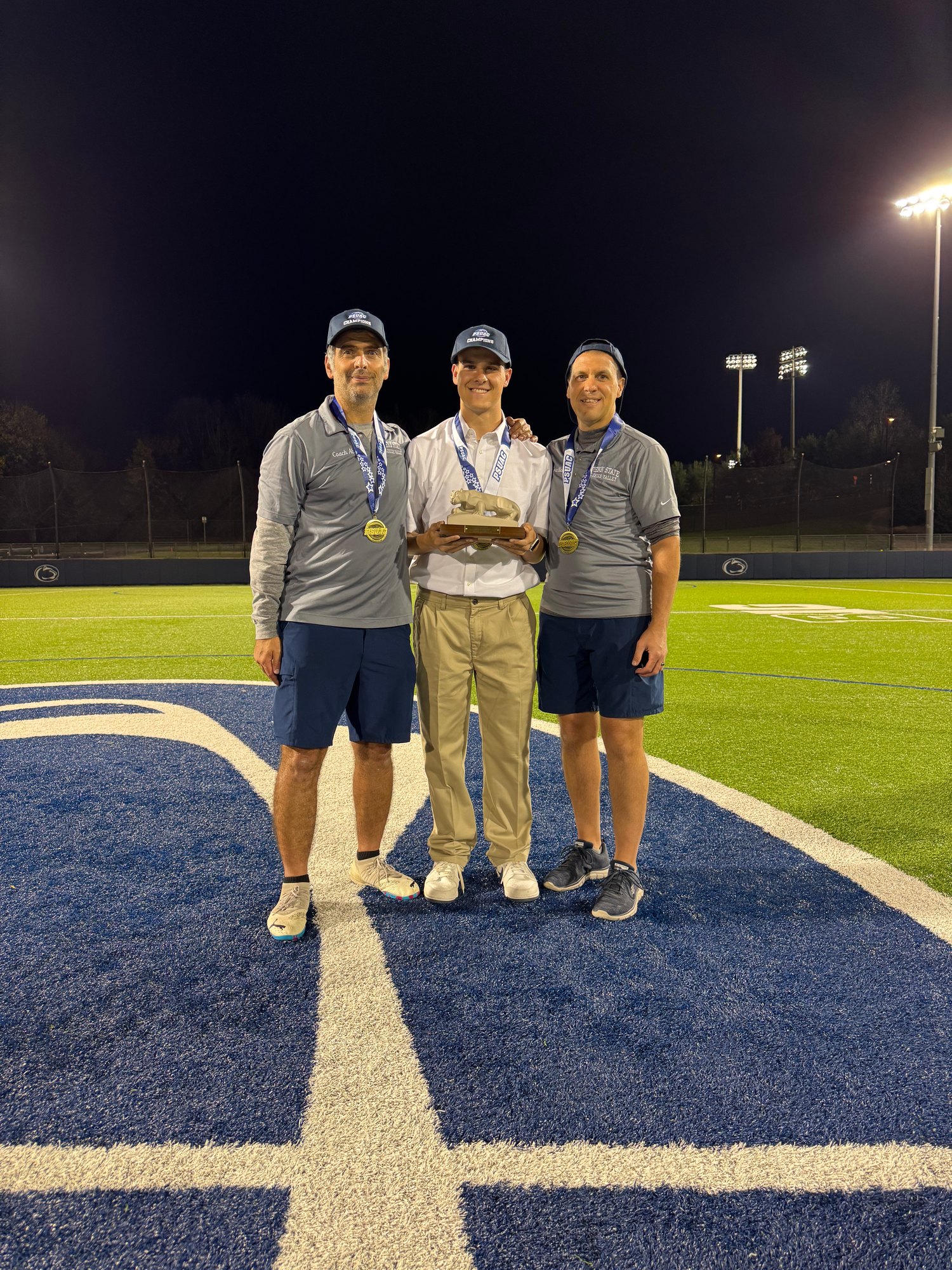 Joseph Draper holds a championship trophy on a Penn State athletics field at night after covering a conference championship